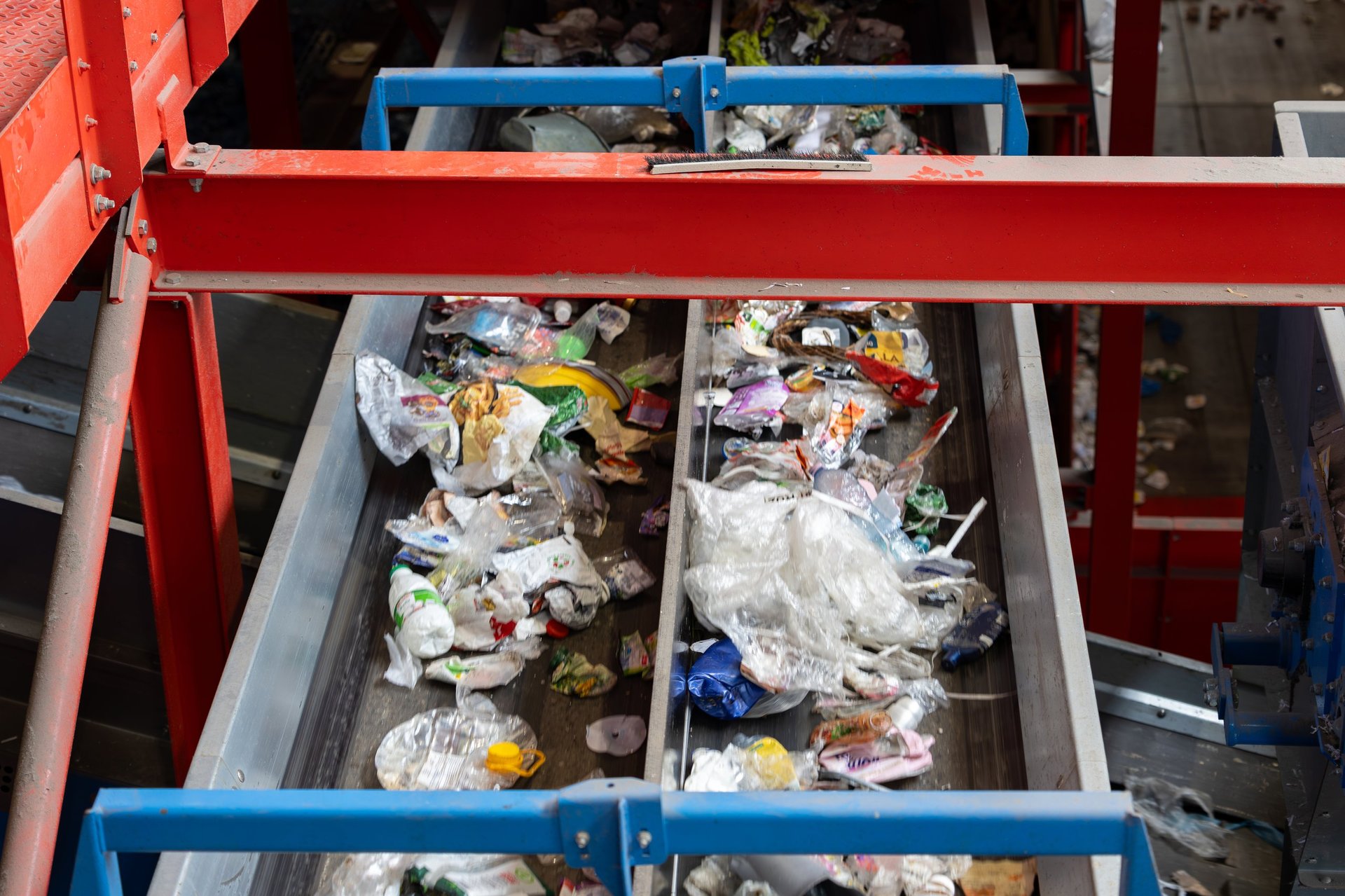 Two parallel conveyor belts transport mixed waste during sorting process inside recycling facility. Plastic bags, packaging, containers and organic residue move along metal channels beneath structural frame. System supports separation and material recovery operations.
